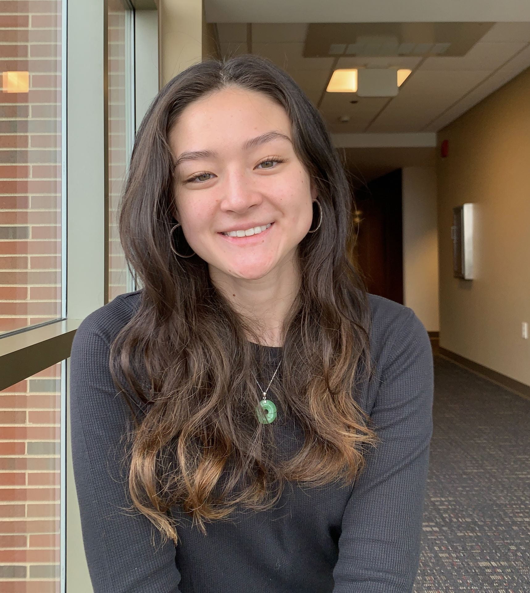 Smiling young person with long wavy hair and a necklace photographed in a hallway